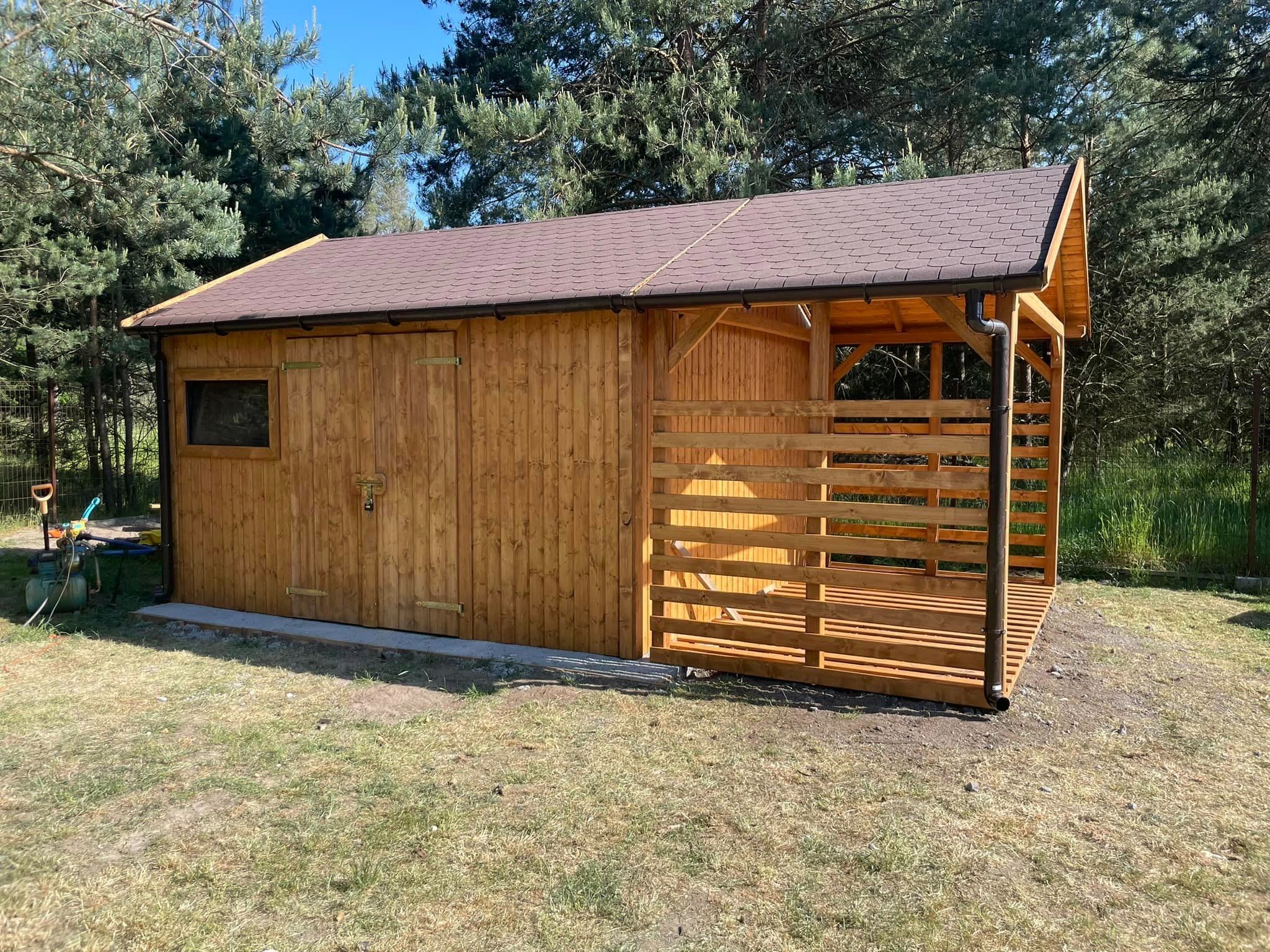 Drewniana konstrukcja z zadaszeniem, łącząca zamkniętą szopę z otwartą przestrzenią tarasową, widoczna na tle drzew