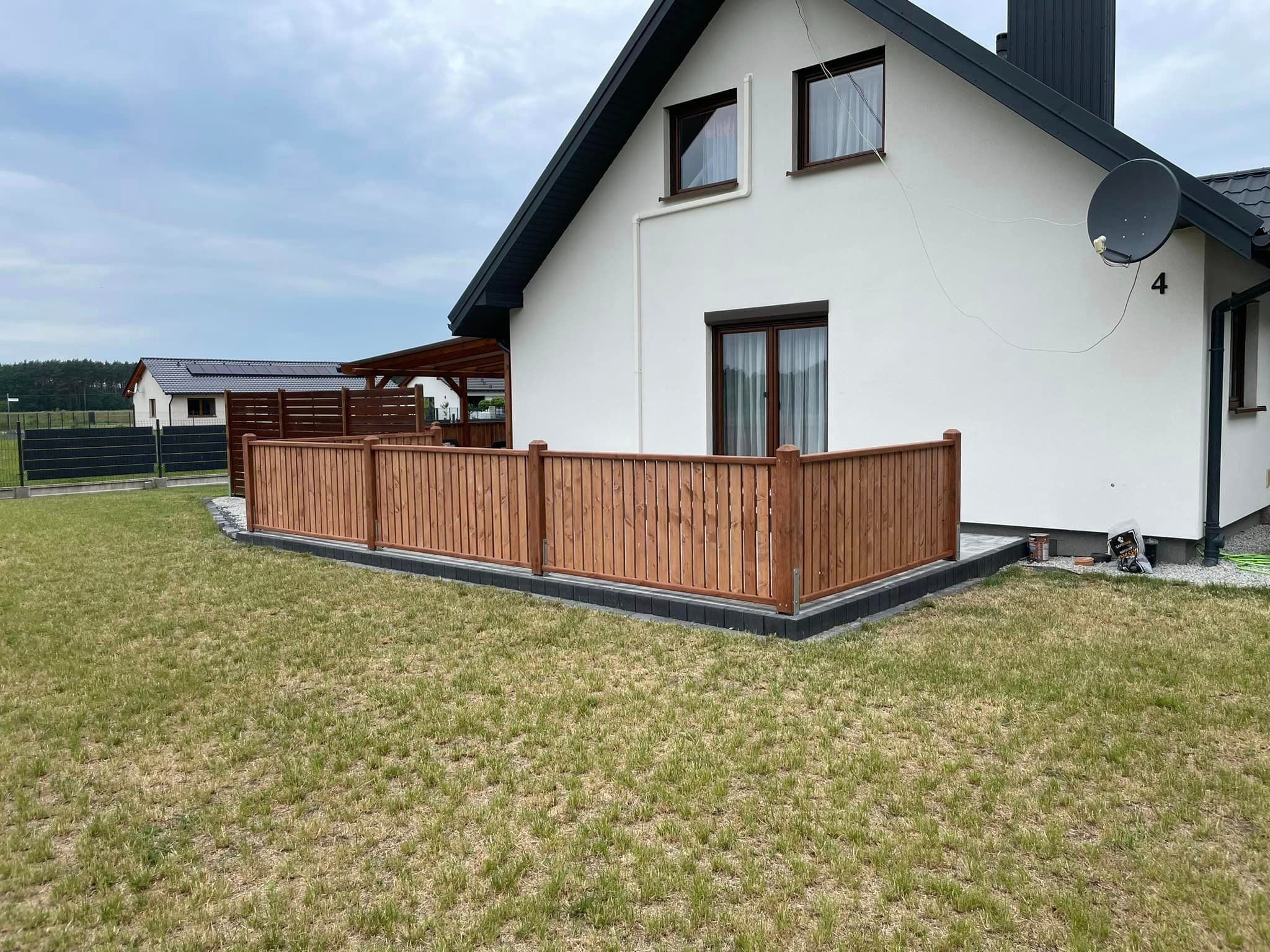 Nowoczesny taras drewniany z niskim ogrodzeniem z pionowych desek, przylegający do białego domu z ciemną stolarką okienną i satelitą na elewacji, na tle zielonego trawnika i sąsiedniego domu...