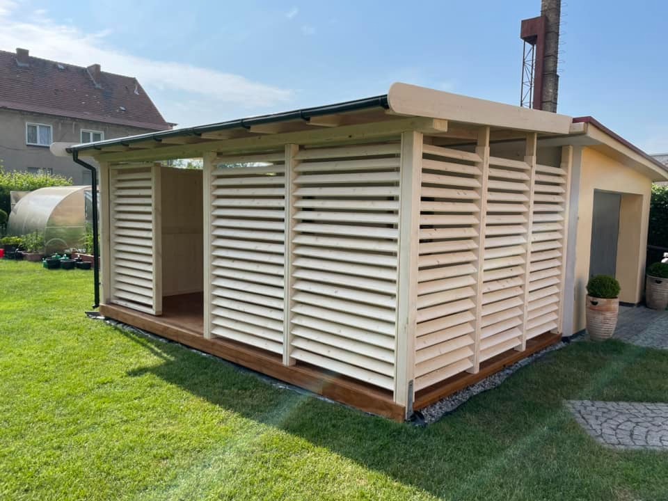 Nowoczesna, jasna konstrukcja z drewnianych żaluzji, tworząca zacienione miejsce na tarasie w ogrodzie, z widocznym budynkiem w tle.