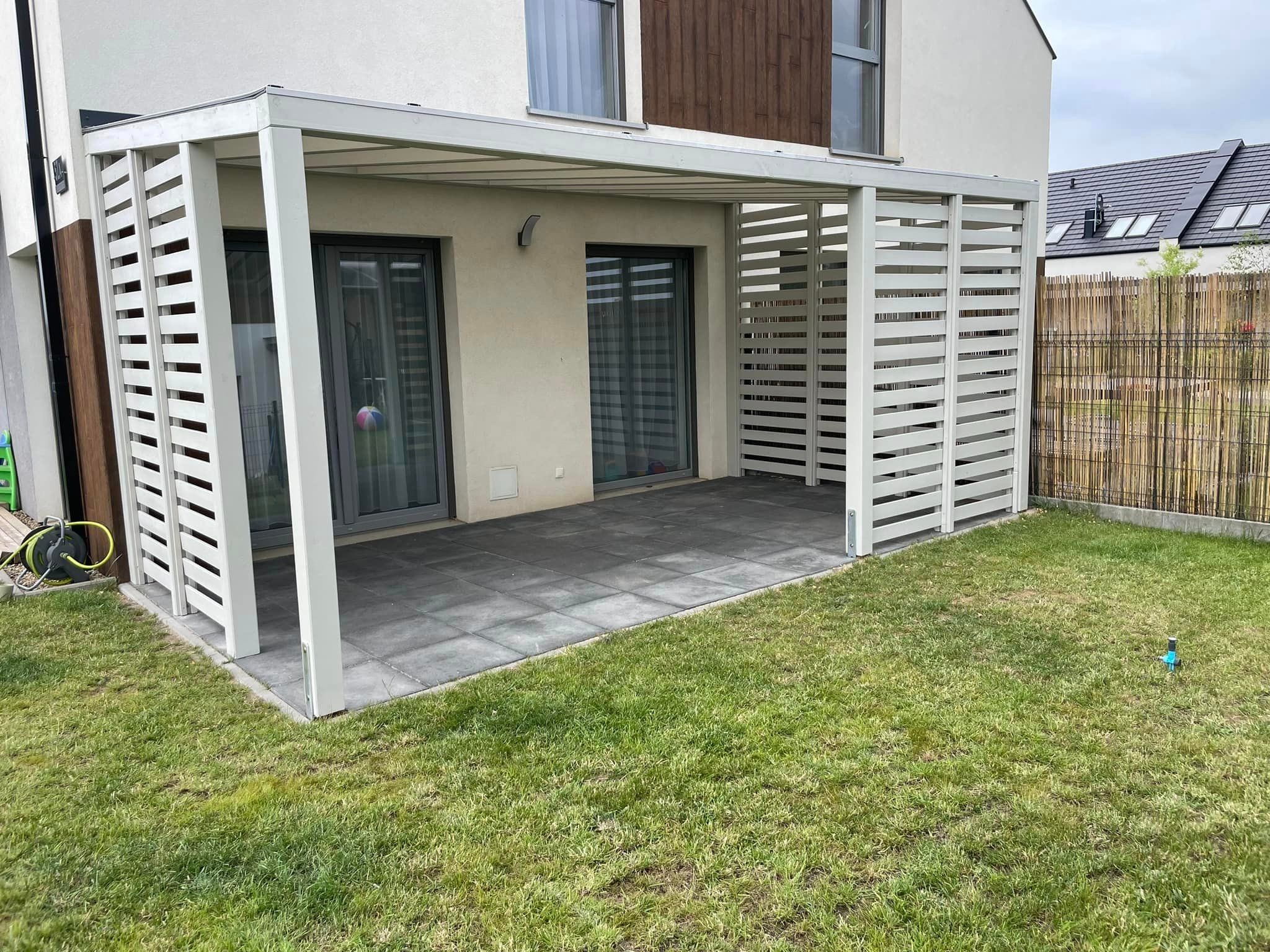 Nowoczesna, biała pergola z drewnianymi lamelami, przylegająca do jasnego budynku z szarymi drzwiami tarasowymi i wyłożonym szarymi płytami tarasem, otoczona zielonym trawnikiem.