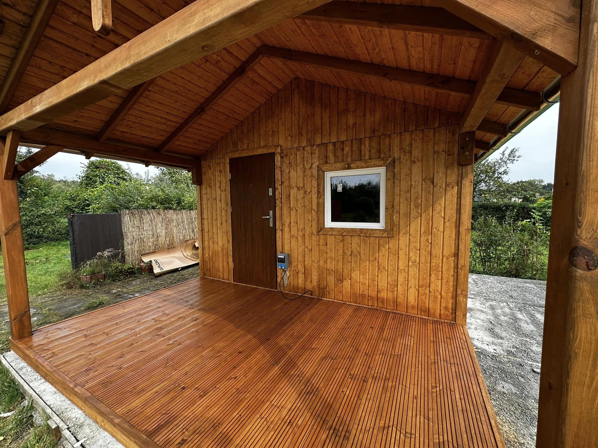 Drewniana konstrukcja z zadaszeniem i tarasem z desek, widoczne drzwi i okno, otoczenie zieleni. Świeżo zaimpregnowana podłoga tarasu. Częściowo widoczny teren wokół konstrukcji.