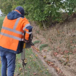 Pracownik w pomarańczowej kamizelce i niebieskich spodniach używa młota pneumatycznego na zewnątrz, w tle drzewa i trawa.