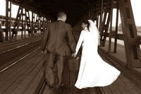 Para młoda, ubrana w tradycyjne stroje ślubne w odcieniach sepii, idzie trzymając się za ręce po drewnianym moście z kratownicową konstrukcją. Ujęcie z tyłu, podkreślające romantyczny charakter...