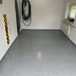 Łukasz Nowak - Szara posadzka żywiczna z kolorowymi drobinkami w garażu, widok na całą powierzchnię, ściany białe, w tle zwinięty wąż ogrodowy i grzejnik pod oknem.