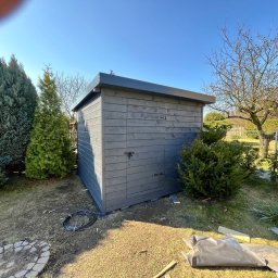 Łukasz Nowak - Szara drewniana szopa ogrodowa z płaskim dachem na tle zieleni i błękitnego nieba. Widoczne elementy montażowe i otoczenie w trakcie prac.