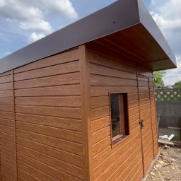 Łukasz Nowak - Nowoczesna, brązowa altana ogrodowa z małym oknem, wykonana z paneli w Ożarowicach. Widoczne ogrodzenie i błękitne niebo w tle.