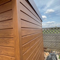 Łukasz Nowak - Brązowy domek narzędziowy z paneli imitujących drewno, widok z boku, z betonowym ażurowym ogrodzeniem w tle i fragmentem nieba z chmurami.