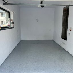 Łukasz Nowak - Szara posadzka żywiczna w garażu z oknem, widoczne worki z materiałami budowlanymi. Ściany białe, surowe wykończenie. Widoczny mechanizm bramy garażowej.