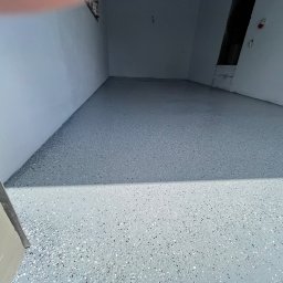 Łukasz Nowak - Szara posadzka żywiczna w garażu lub pomieszczeniu gospodarczym, z bliska widoczne drobne, kolorowe wtrącenia w żywicy. Ściany pomalowane na jasny odcień szarości.