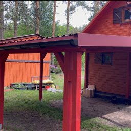 Łukasz Nowak - Czerwona, drewniana pergola przy domu z drewnianą elewacją. Widoczne drzewa w tle. Pergola z metalowym dachem i drewnianymi słupami. Ułożona kostka brukowa.