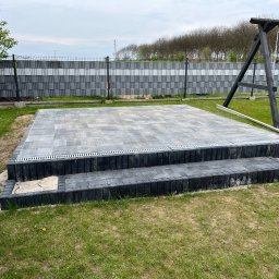 Łukasz Nowak - Nowy taras z szarej kostki brukowej, dwupoziomowy, z odpływem liniowym, otoczony zielonym trawnikiem i nowoczesnym ogrodzeniem. W tle huśtawka.