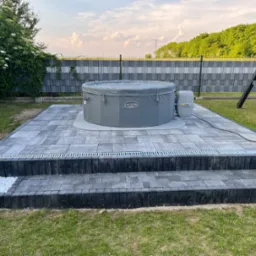 Szary, nadmuchiwany basen Lay-Z-Spa na dwupoziomowym tarasie z szarej kostki brukowej, z odpływem liniowym i dekoracyjnymi białymi kamieniami. W tle zielona roślinność i ogrodzenie.