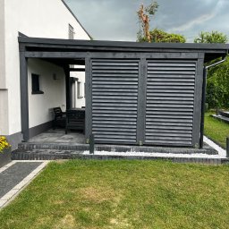 Łukasz Nowak - Nowoczesna pergola z szarymi lamelami i białymi kamieniami ozdobnymi na tle zielonego trawnika, w tle dom z białymi ścianami i szarymi akcentami.