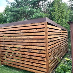 Łukasz Nowak - Drewniana pergola z brązowym, metalowym dachem na zielonym trawniku, widok z boku. W tle drzewa i częściowo widoczny stół z krzesłami.