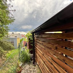Łukasz Nowak - Drewniana konstrukcja z brązowych desek, widok z dołu na zewnątrz, z fragmentem rynny i zielenią w tle, pod pochmurnym niebem. Elementy wykończenia ogrodu.