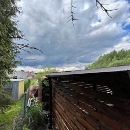 Łukasz Nowak - Drewniana pergola z brązowym dachem i rynną, widoczna z boku, z zielenią w tle i pochmurnym niebem. Elementy ogrodu, drzewa i budynki w oddali.