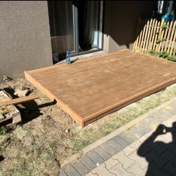 Łukasz Nowak - Wykończony taras drewniany przy domu z butelką wody. Widoczne deski tarasowe, paleta i cień osoby fotografującej. Częściowo widoczny płot i trawa.