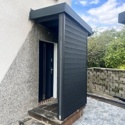 Łukasz Nowak - Zadaszenie nad wejściem do domu z czarnego kompozytu, dopasowane kolorystycznie do drzwi. Obok ściana z szarego tynku strukturalnego i fragment kostki brukowej.