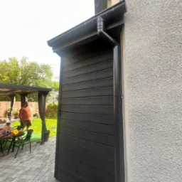 Ciemna okładzina elewacyjna z desek kompozytowych, widoczny fragment orynnowania i ściana budynku o szorstkiej fakturze. W tle fragment tarasu i pergola.