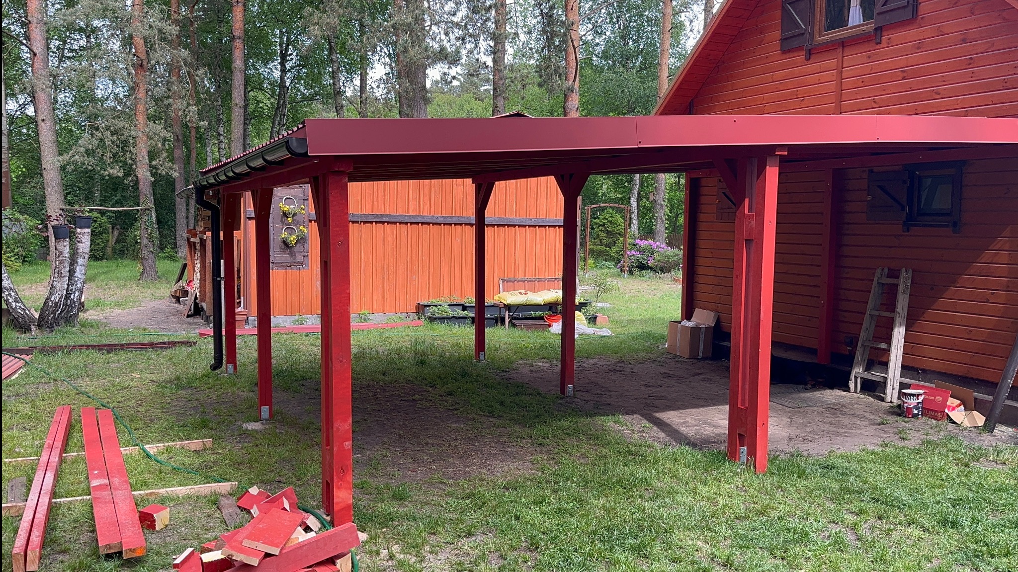 Czerwona pergola przy domu w lesie, w trakcie montażu. Widoczne elementy konstrukcyjne i narzędzia. Naturalne oświetlenie, leśne otoczenie.