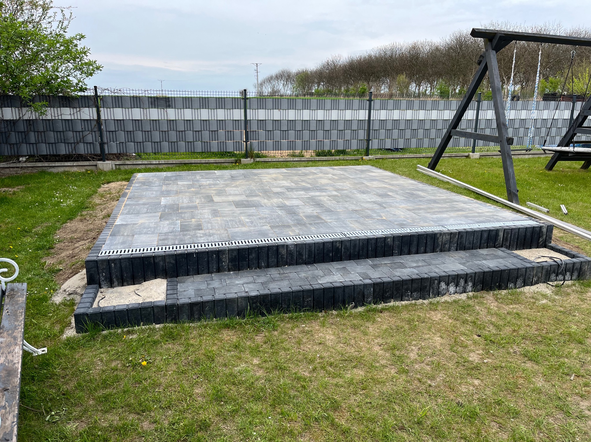 Nowy taras z szarej kostki brukowej, dwupoziomowy, z odpływem liniowym, otoczony zielonym trawnikiem i nowoczesnym ogrodzeniem. W tle huśtawka.