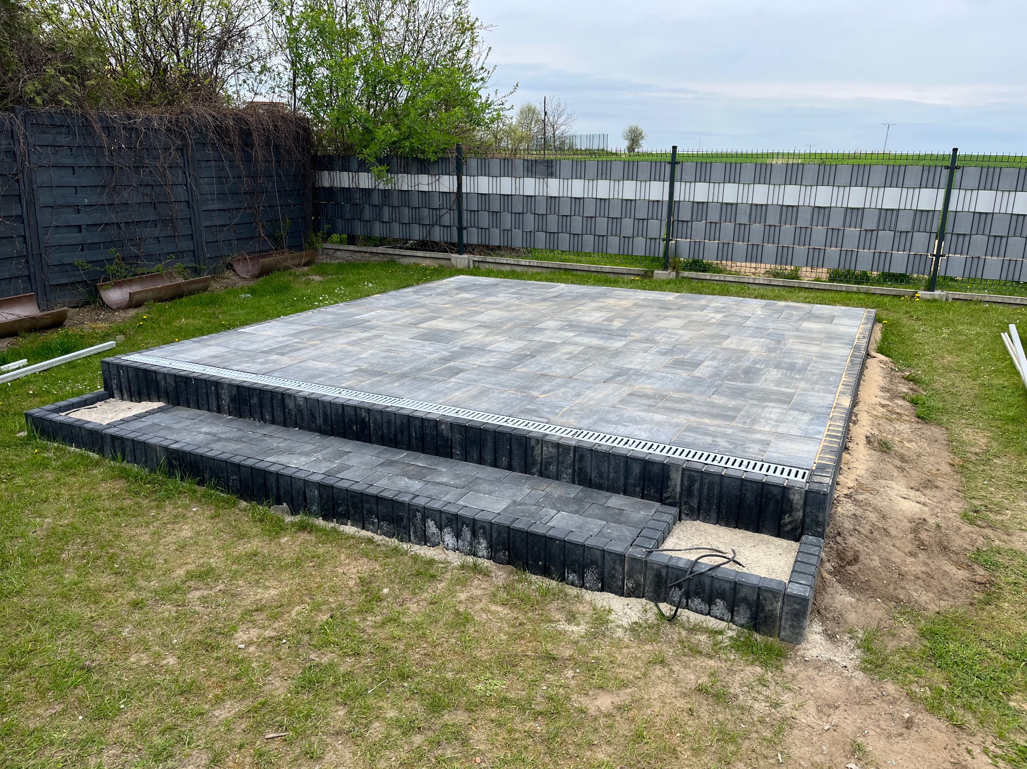 Wykończony taras z kostki brukowej w ogrodzie, z dwoma stopniami i odpływem liniowym. Widoczne ogrodzenie panelowe i fragment zieleni.