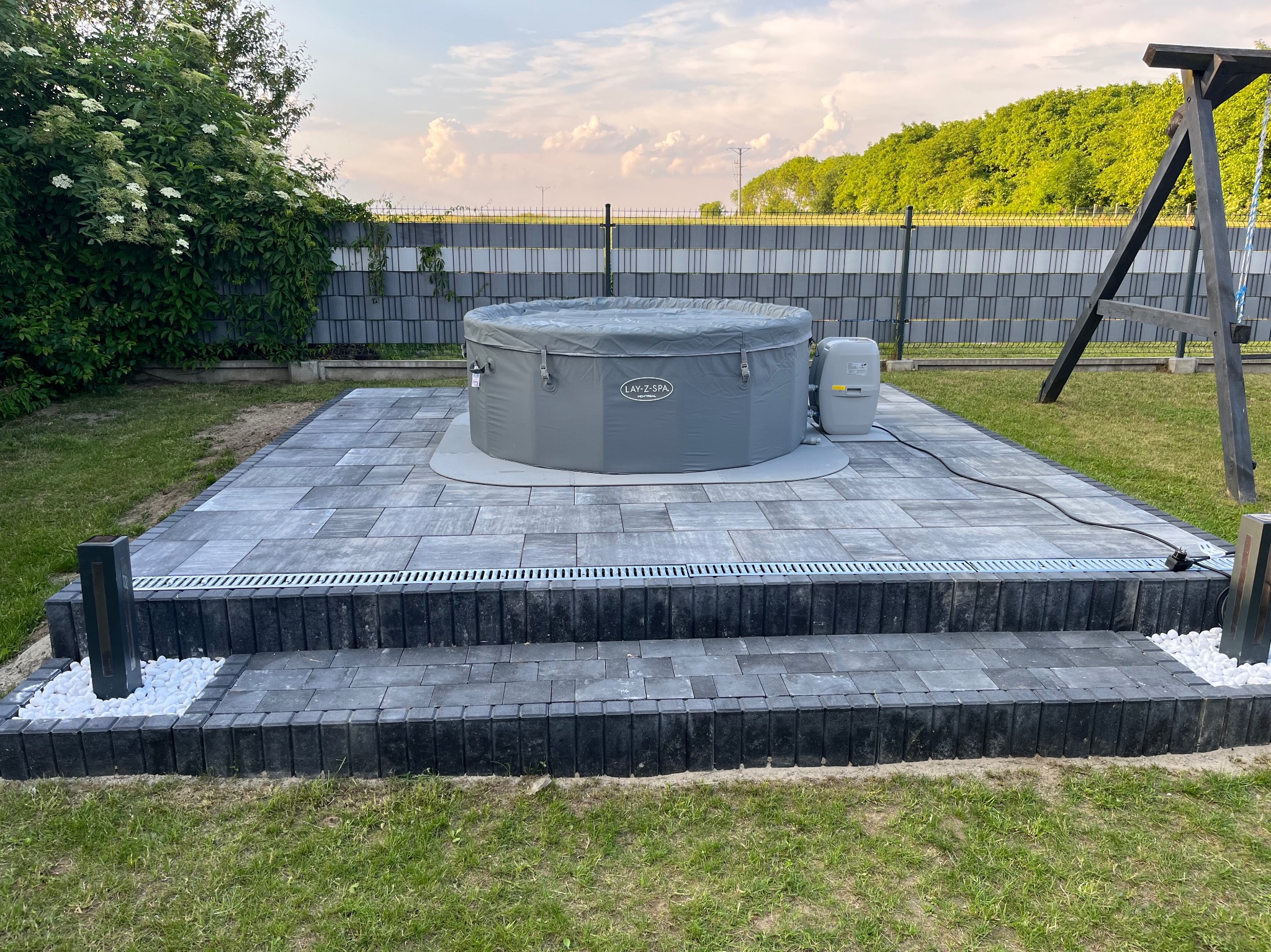 Szary, nadmuchiwany basen Lay-Z-Spa na dwupoziomowym tarasie z szarej kostki brukowej, z odpływem liniowym i dekoracyjnymi białymi kamieniami. W tle zielona roślinność i ogrodzenie.
