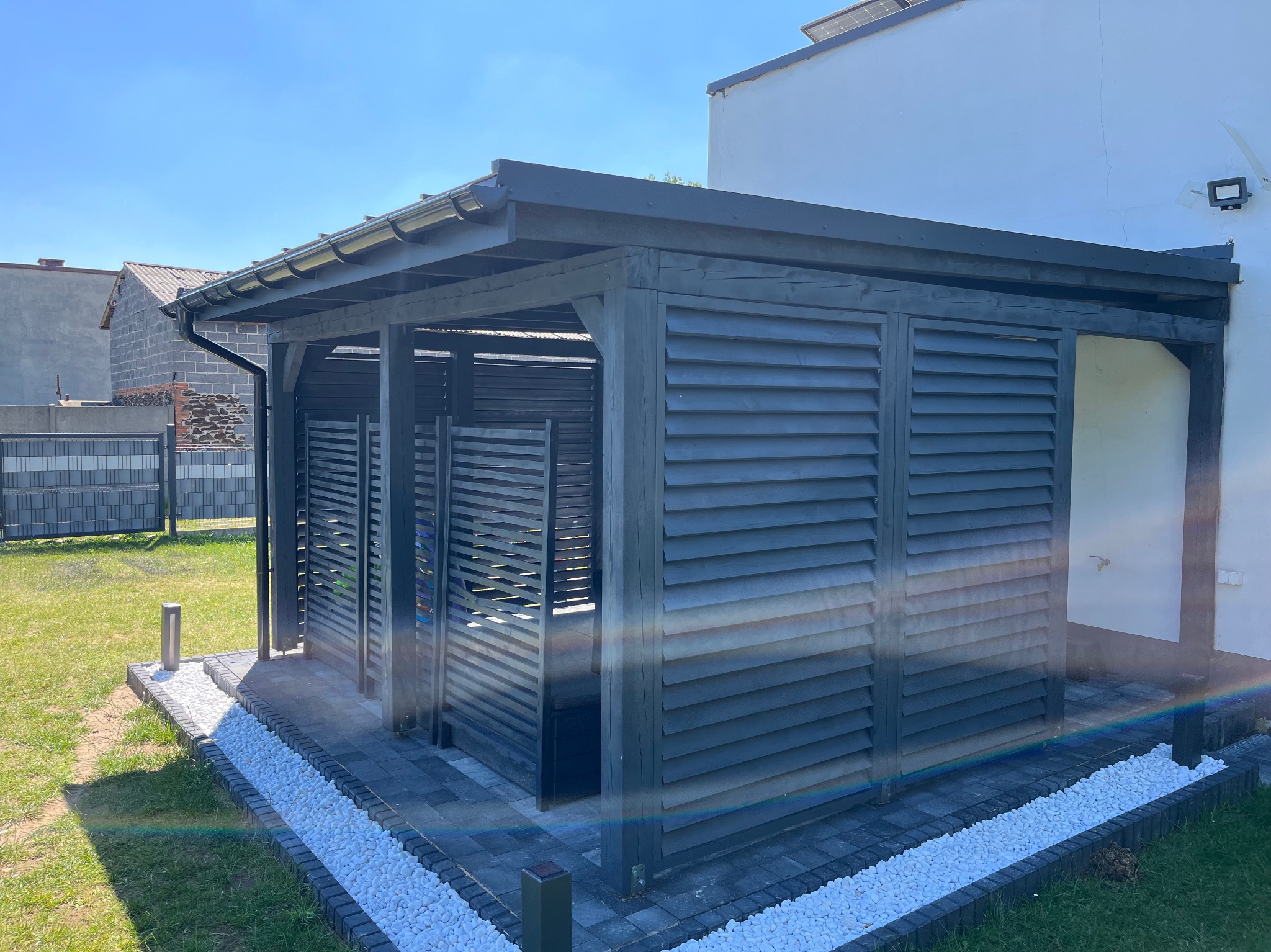Szara pergola z żaluzjowymi ścianami, obłożona białymi kamykami, na tle zielonego trawnika i białego budynku w słoneczny dzień.