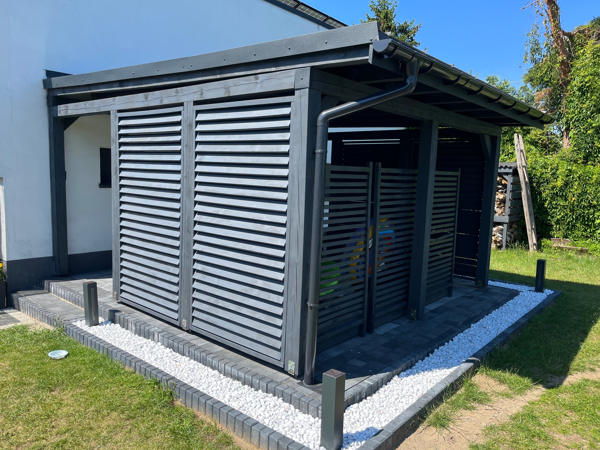 Nowoczesna, szara pergola drewniana z żaluzjowymi ścianami, obłożona białym kamieniem, na tle zielonej trawy i budynku. Widoczny system rynnowy.