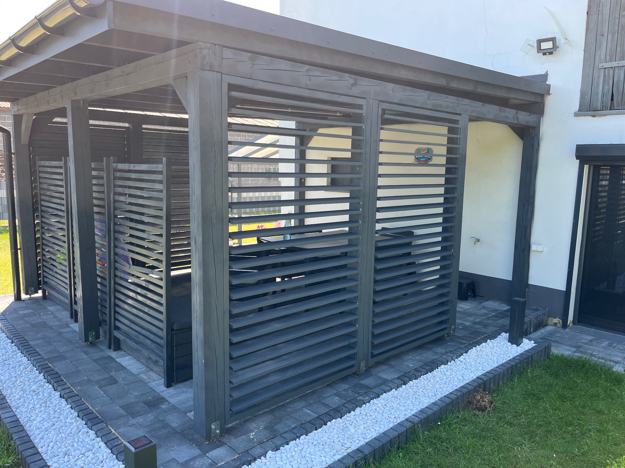 Nowoczesna pergola z ciemnego drewna z poziomymi listwami, na tle białego budynku. Ułożona kostka brukowa i białe kamienie dekoracyjne. Widok z zewnątrz.