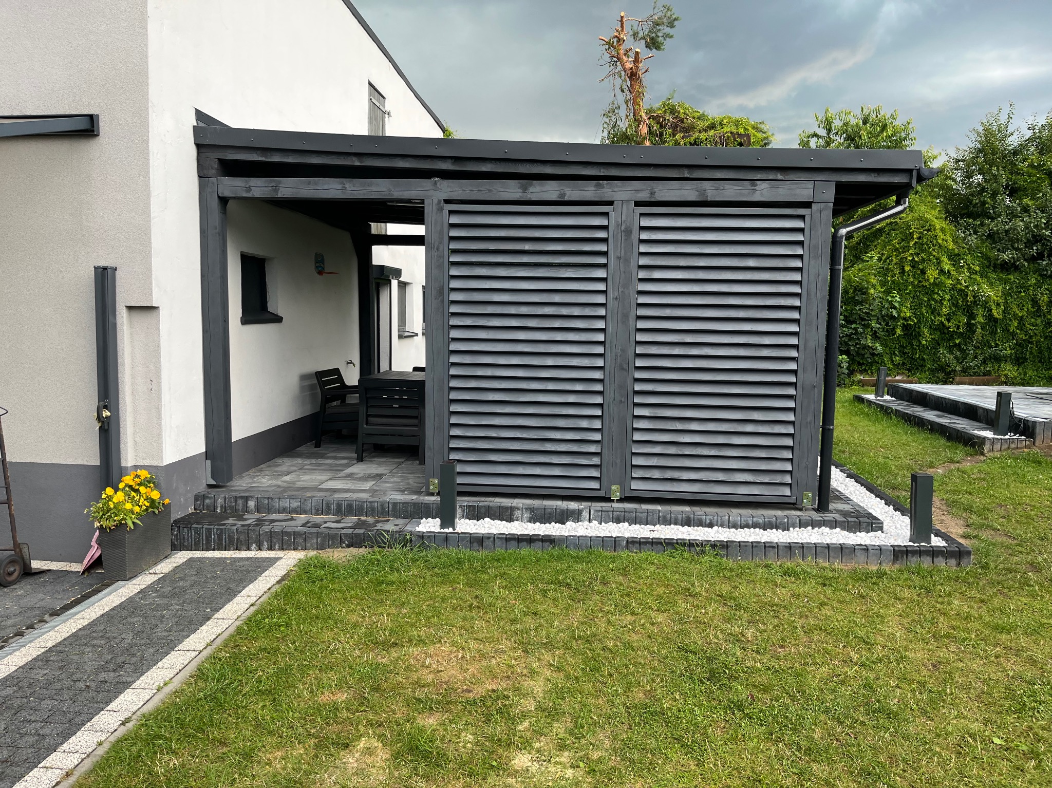 Nowoczesna pergola z szarymi lamelami i białymi kamieniami ozdobnymi na tle zielonego trawnika, w tle dom z białymi ścianami i szarymi akcentami.