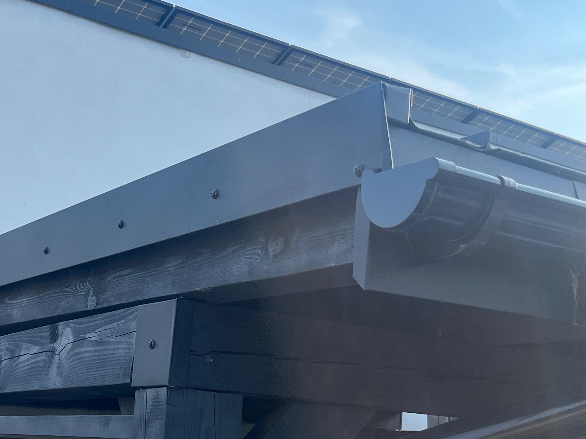 Detal zadaszenia: czarna drewniana konstrukcja pergoli z metalowym orynnowaniem na tle błękitnego nieba i fragmentu ściany budynku z panelami fotowoltaicznymi.