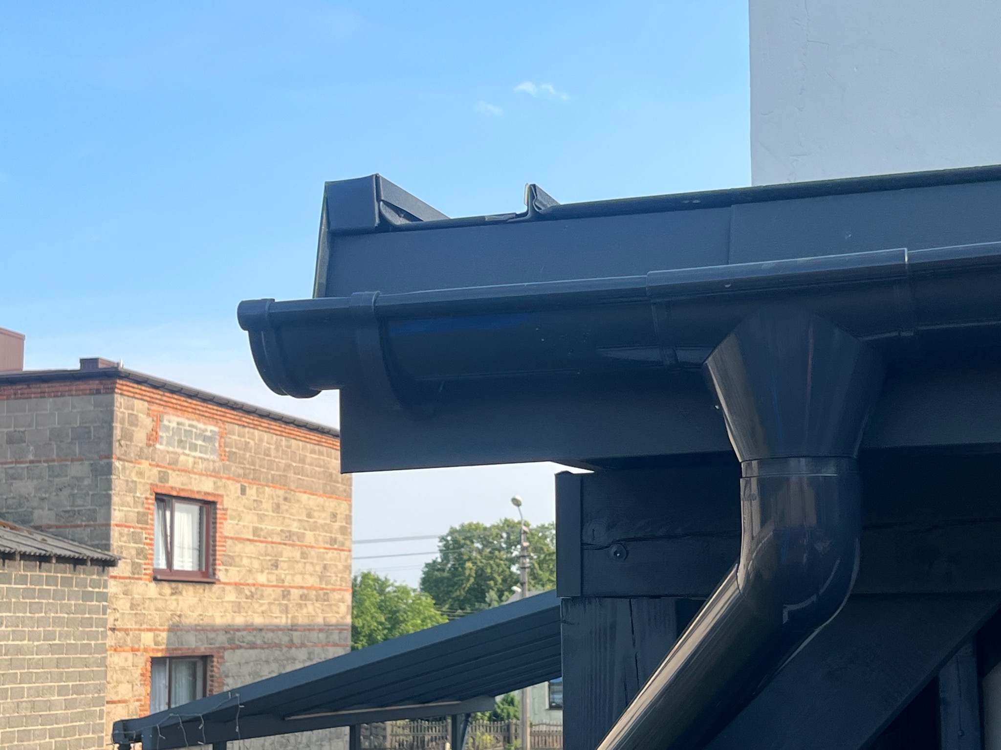 Szczegółowe ujęcie nowej, czarnej rynny dachowej z odpływem, na tle błękitnego nieba i budynku z cegły. Widoczne elementy konstrukcji dachu i orynnowania.