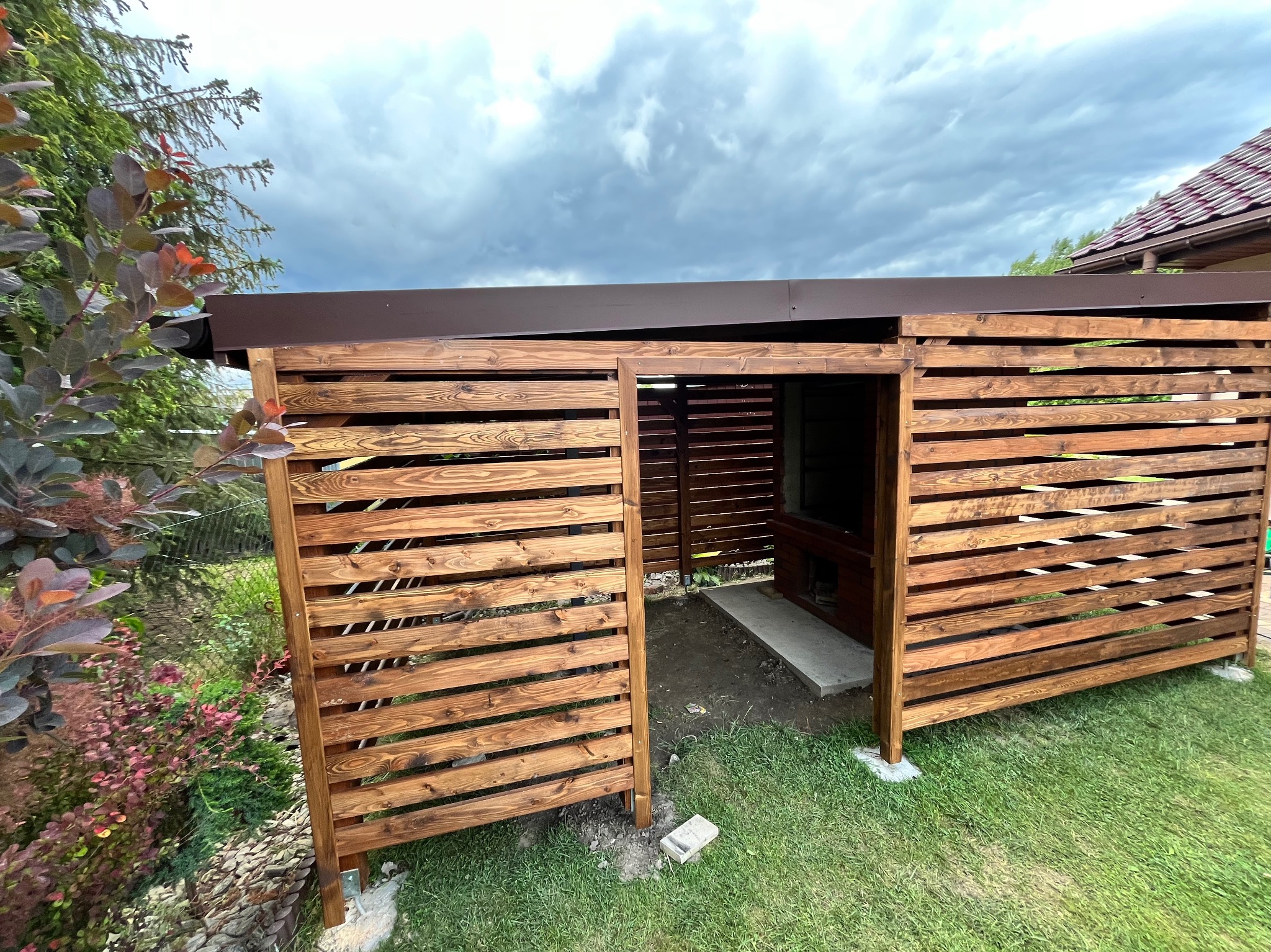 Drewniana pergola z brązowym dachem, lamelowa konstrukcja, widok z zewnątrz na tle zieleni i pochmurnego nieba. Elementy roślinne w lewym dolnym rogu.