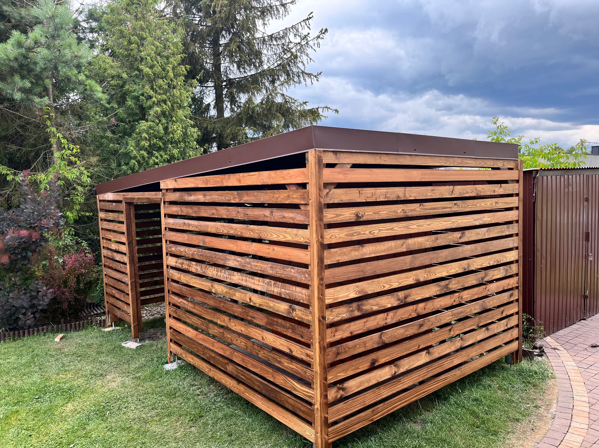 Drewniana pergola z brązowym dachem na trawniku, widok z boku. Konstrukcja z poziomych desek, tło stanowią drzewa i fragment metalowego ogrodzenia.