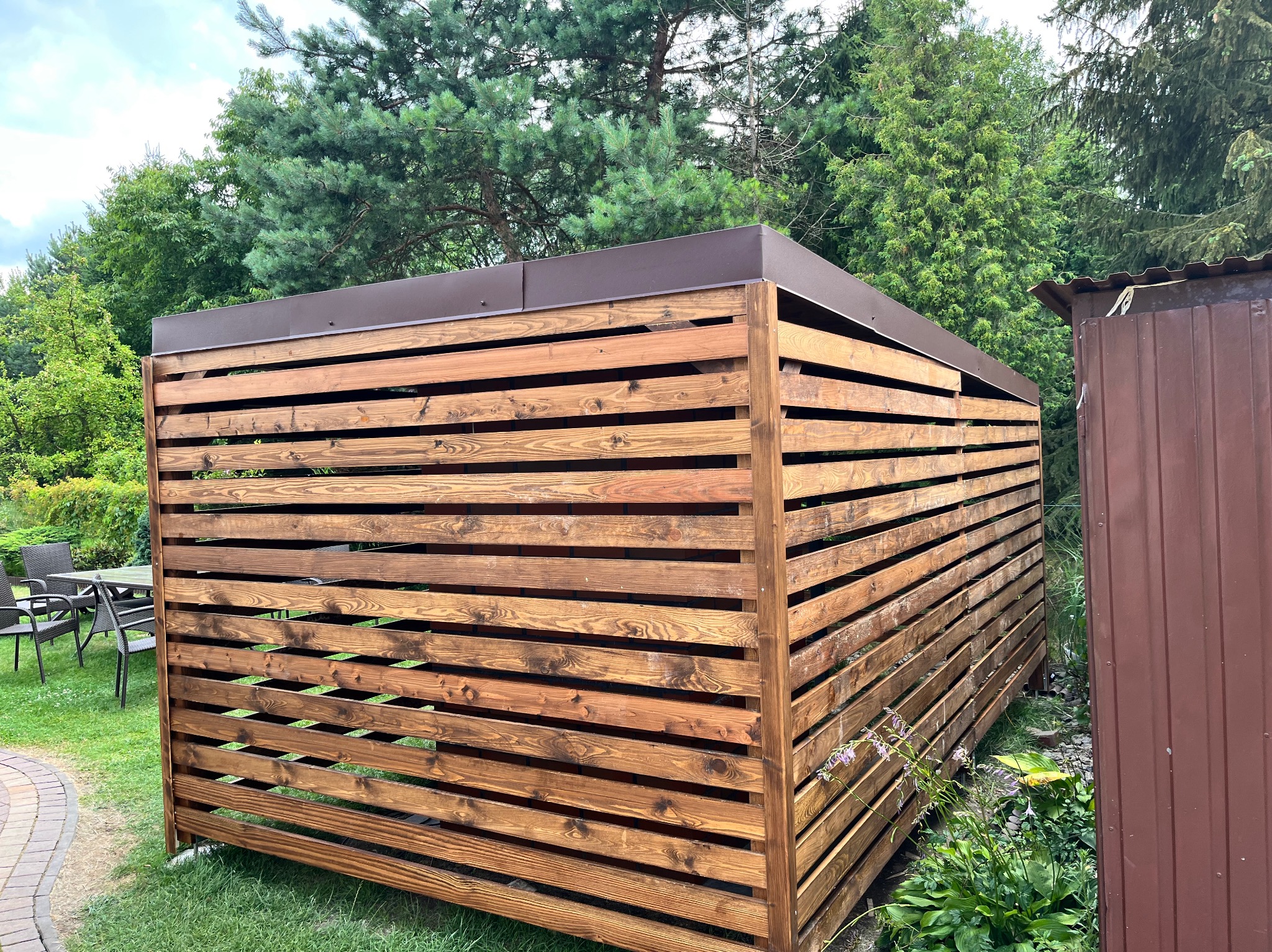 Drewniana pergola z brązowym, metalowym dachem na zielonym trawniku, widok z boku. W tle drzewa i częściowo widoczny stół z krzesłami.