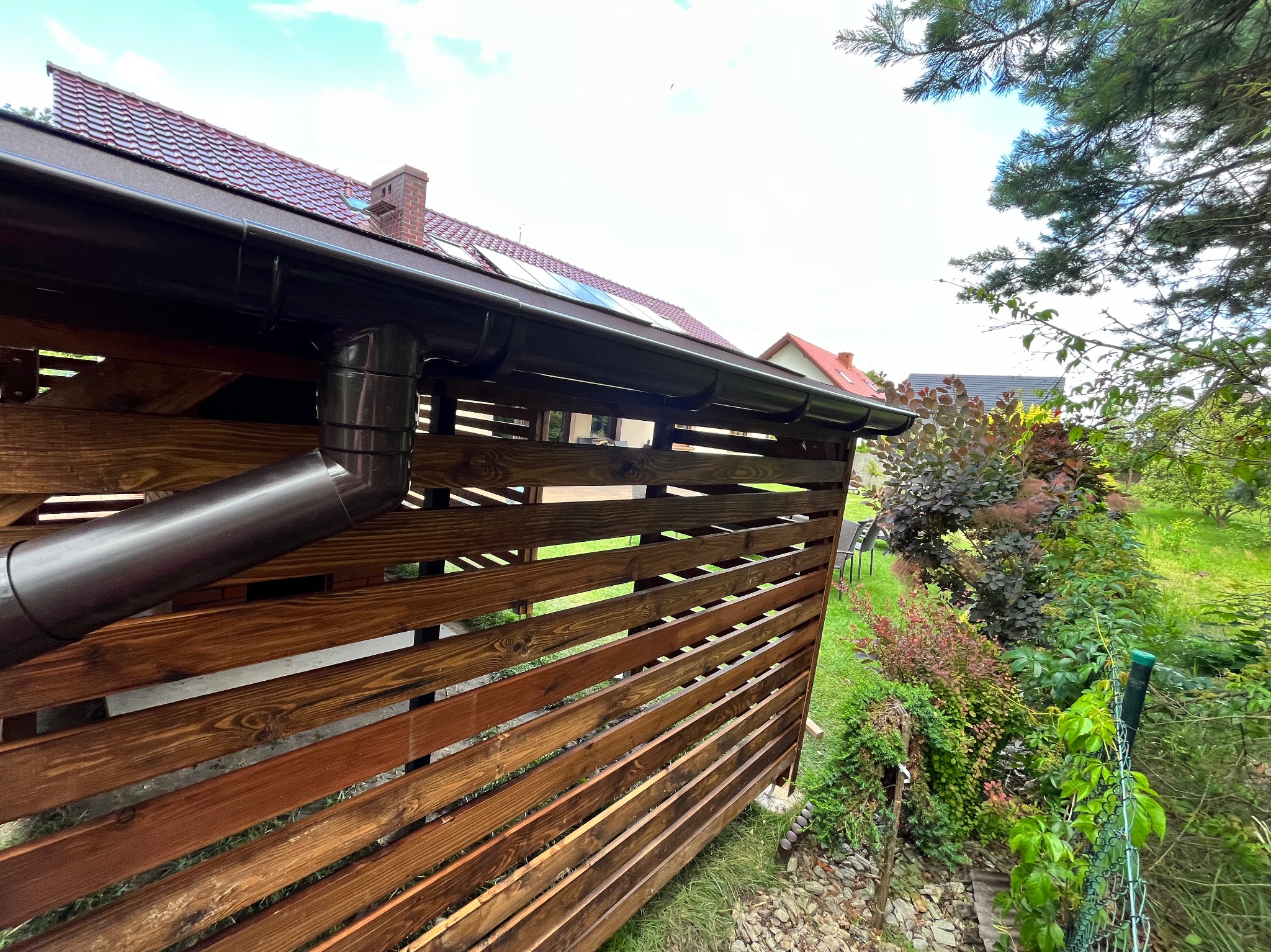 Drewniana pergola z brązowymi rynnami w ogrodzie, widok z dołu. Uwagę przyciąga starannie wykonana konstrukcja i dopasowane kolorystycznie orynnowanie.