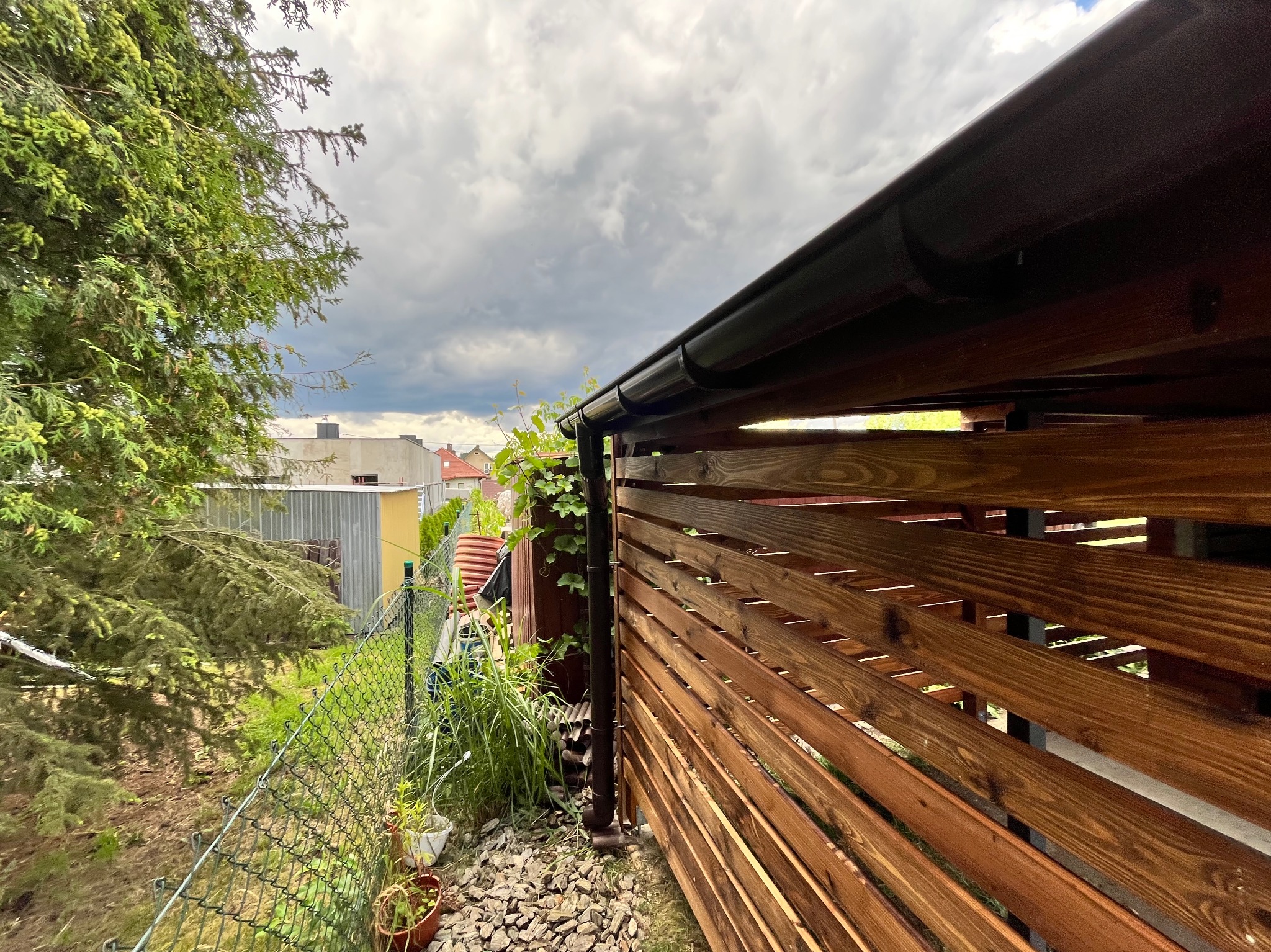 Drewniana konstrukcja z brązowych desek, widok z dołu na zewnątrz, z fragmentem rynny i zielenią w tle, pod pochmurnym niebem. Elementy wykończenia ogrodu.