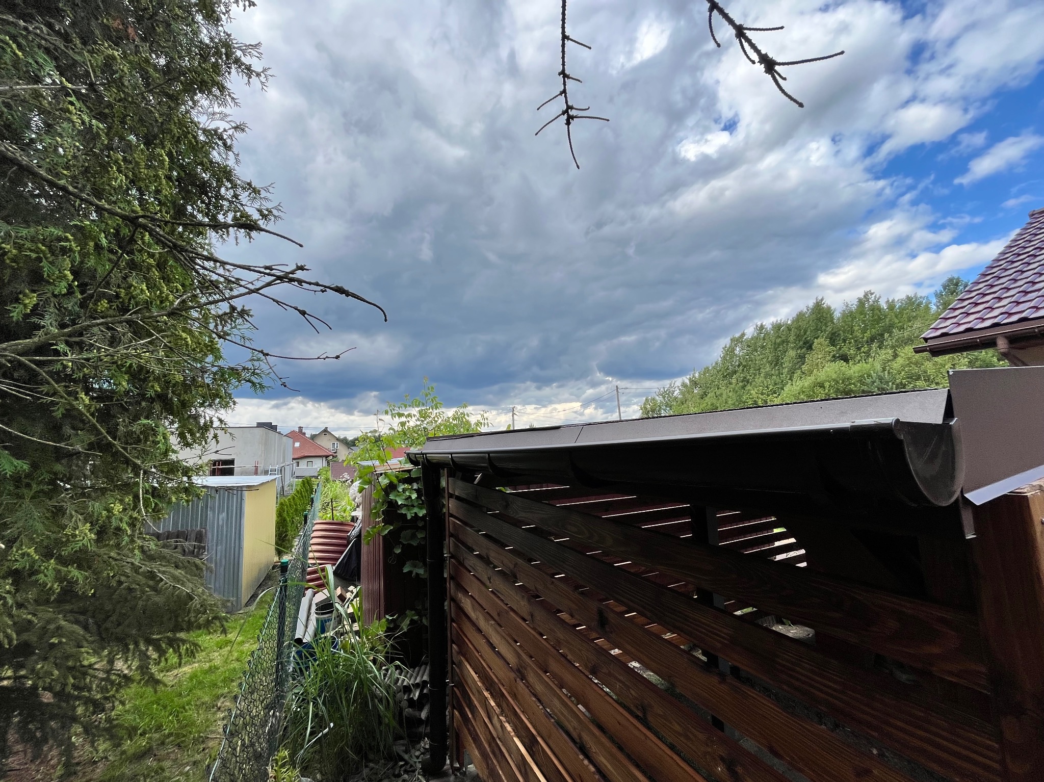 Drewniana pergola z brązowym dachem i rynną, widoczna z boku, z zielenią w tle i pochmurnym niebem. Elementy ogrodu, drzewa i budynki w oddali.