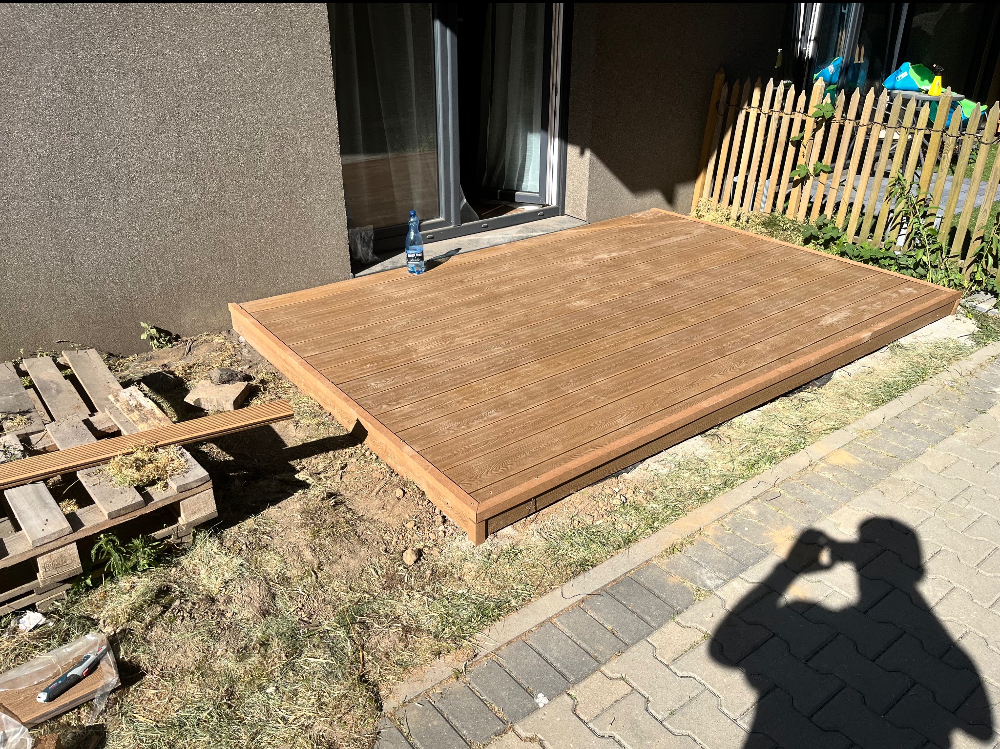 Wykończony taras drewniany przy domu z butelką wody. Widoczne deski tarasowe, paleta i cień osoby fotografującej. Częściowo widoczny płot i trawa.