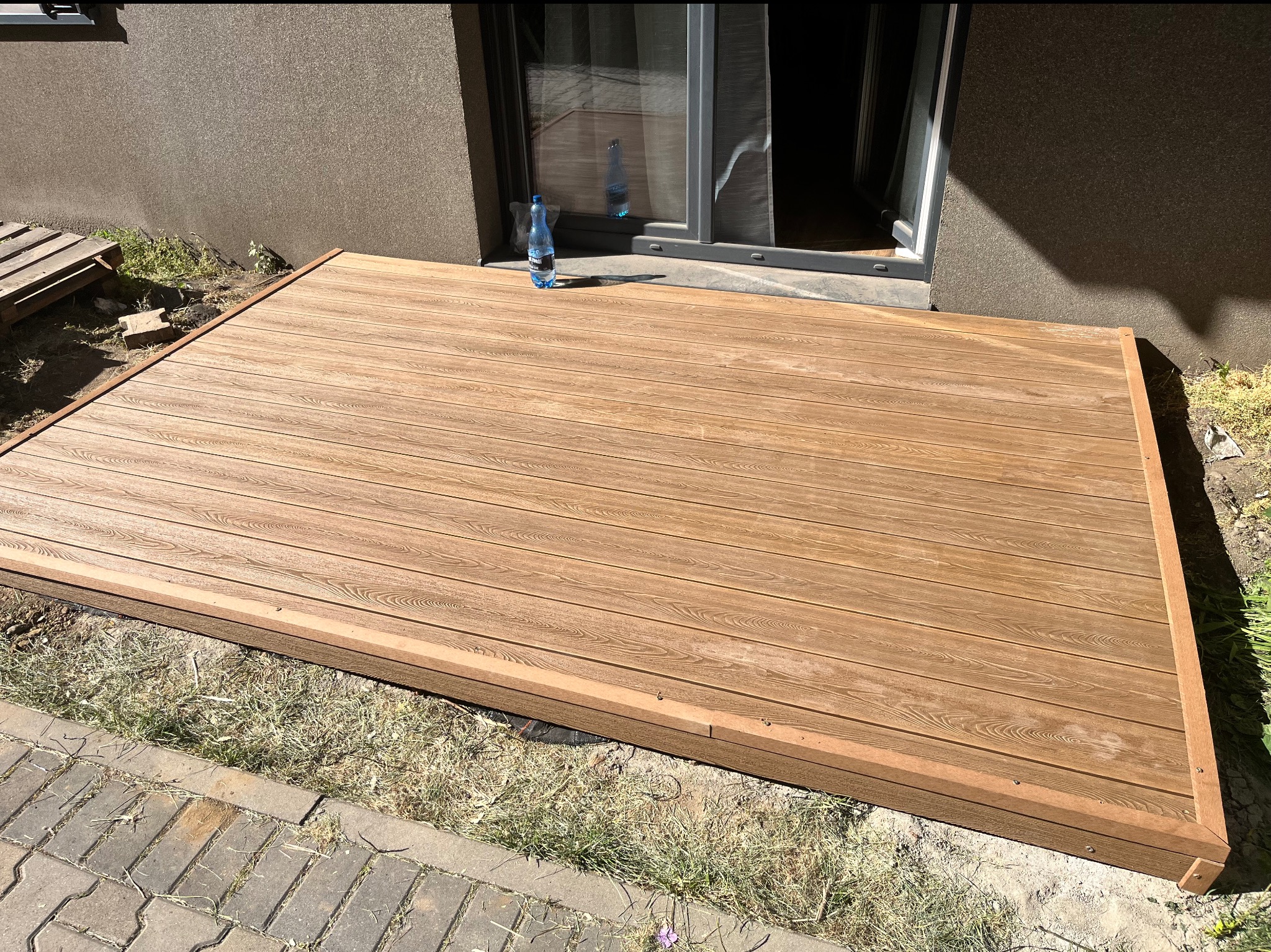 Nowy taras kompozytowy w odcieniach brązu przylega do ściany budynku. Widoczna butelka wody. Estetyczne wykończenie obrzeży. Trawa i kostka brukowa w tle.