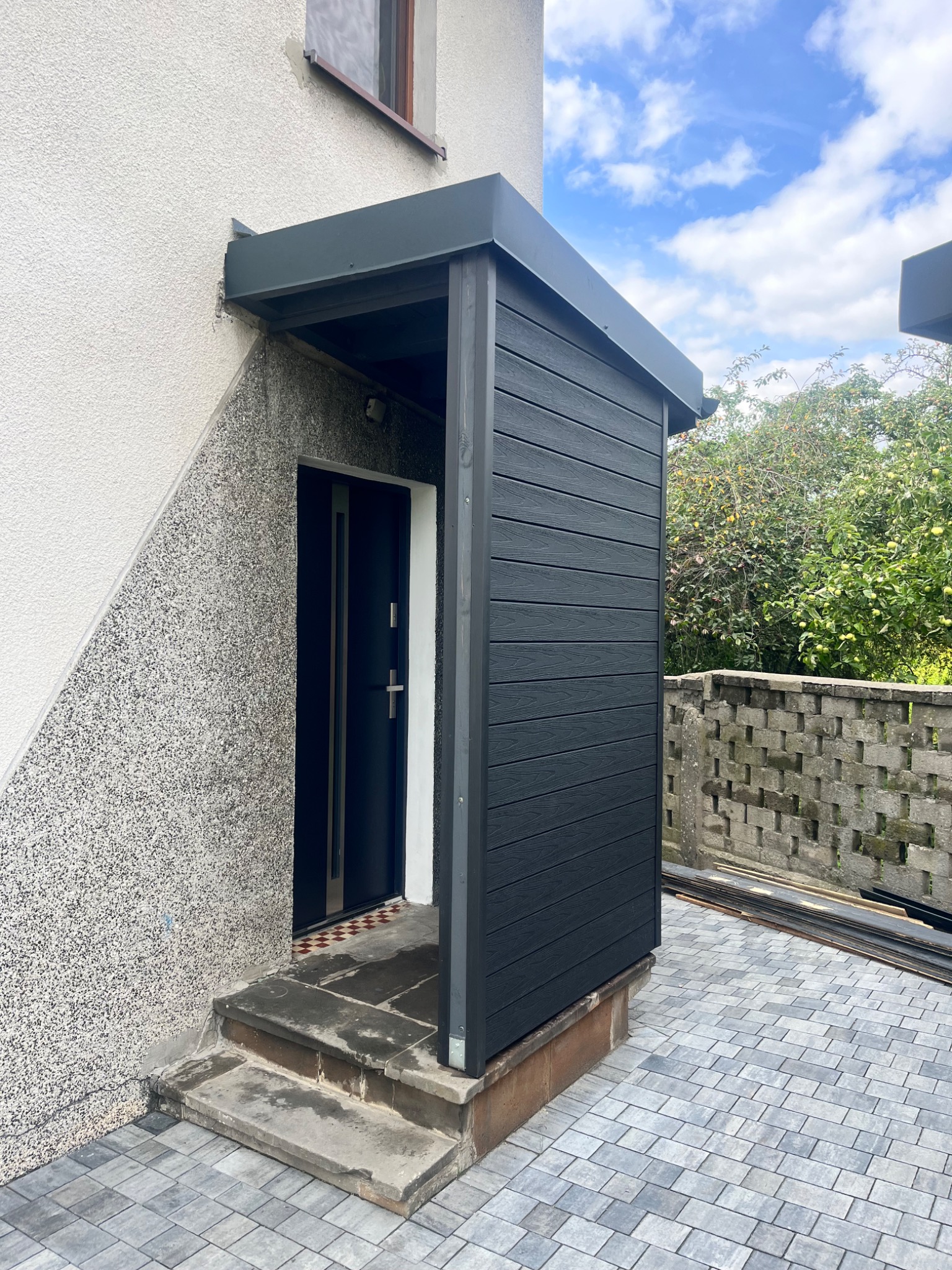 Zadaszenie nad wejściem do domu z czarnego kompozytu, dopasowane kolorystycznie do drzwi. Obok ściana z szarego tynku strukturalnego i fragment kostki brukowej.