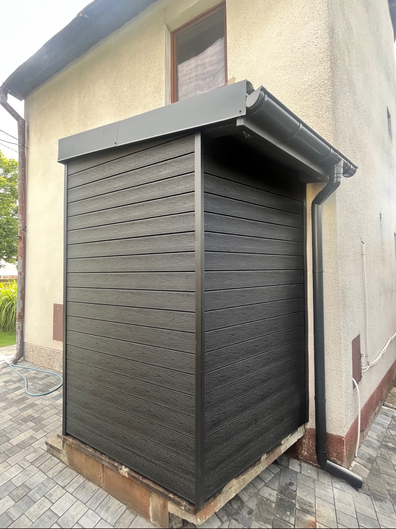 Nowoczesna zabudowa tarasowa z desek kompozytowych w kolorze grafitowym, przylegająca do ściany budynku. Widoczne orynnowanie i kostka brukowa.