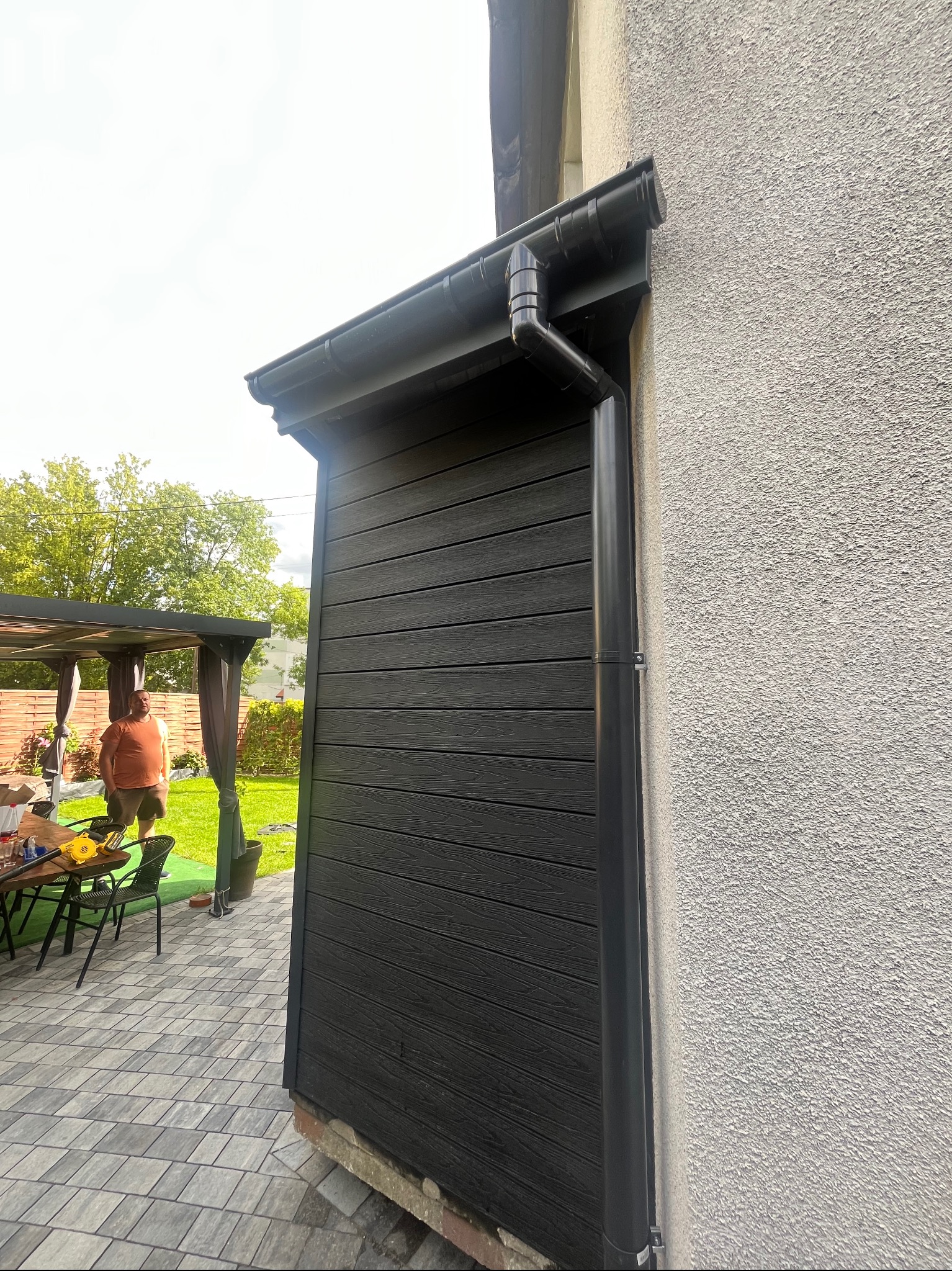Ciemna okładzina elewacyjna z desek kompozytowych, widoczny fragment orynnowania i ściana budynku o szorstkiej fakturze. W tle fragment tarasu i pergola.