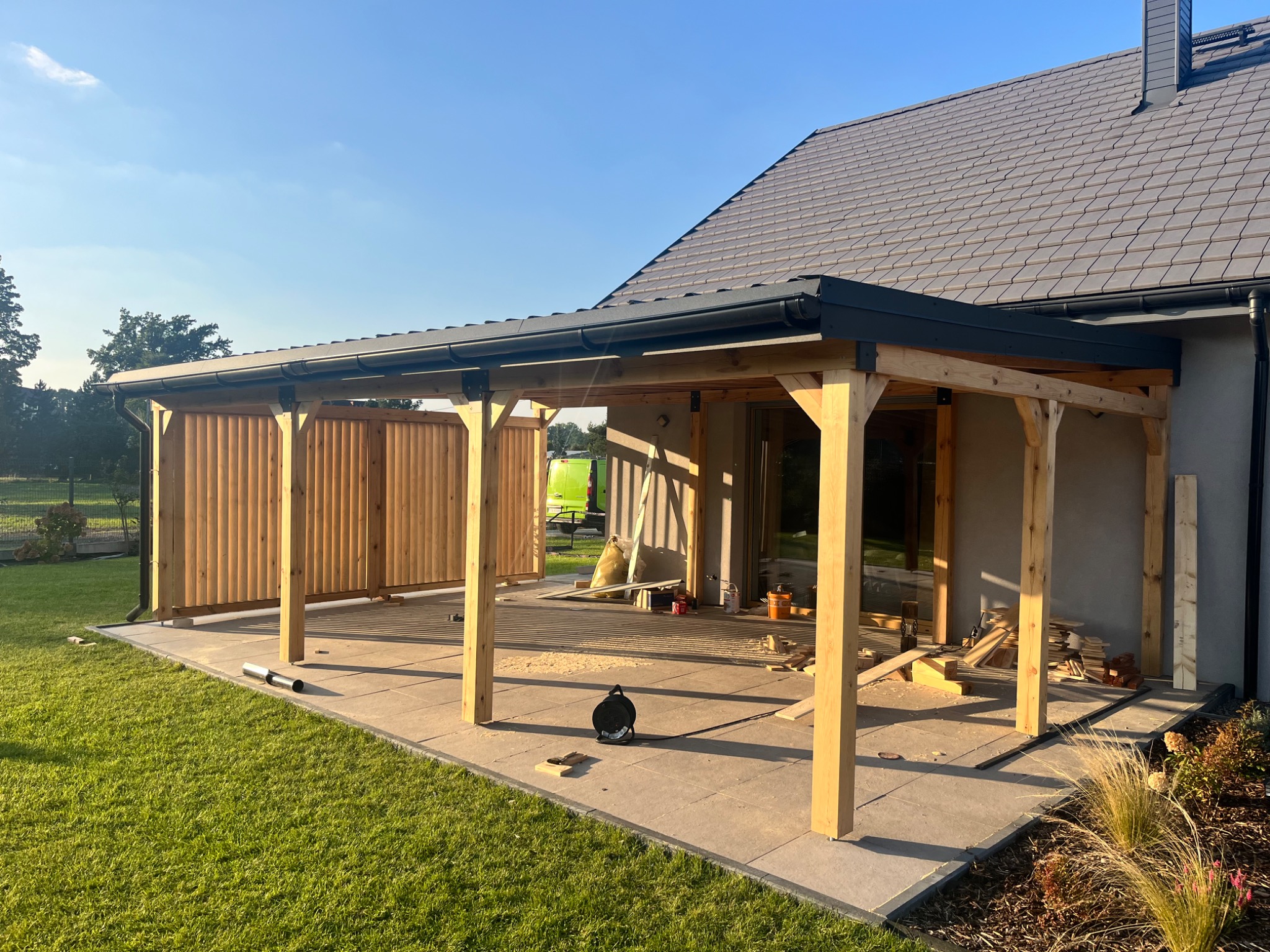 Drewniana pergola w trakcie budowy, z widocznymi słupami i zadaszeniem, na tle domu z szarą elewacją i zielonym trawnikiem w słoneczny dzień. Widoczne narzędzia i materiały budowlane.
