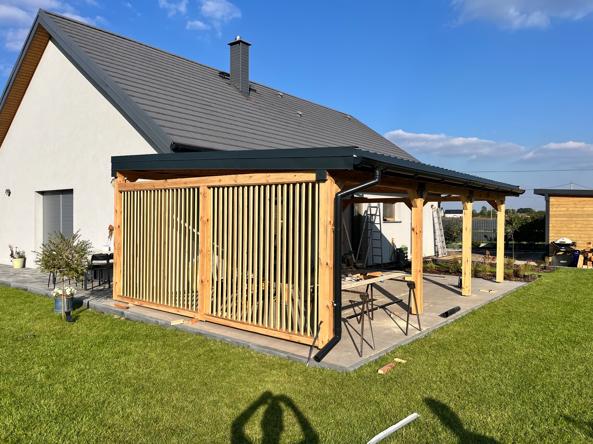 Drewniana pergola z lamelami w ogrodzie, w trakcie montażu. Widoczny fragment domu z szarą dachówką i zielony trawnik. Słoneczny dzień.