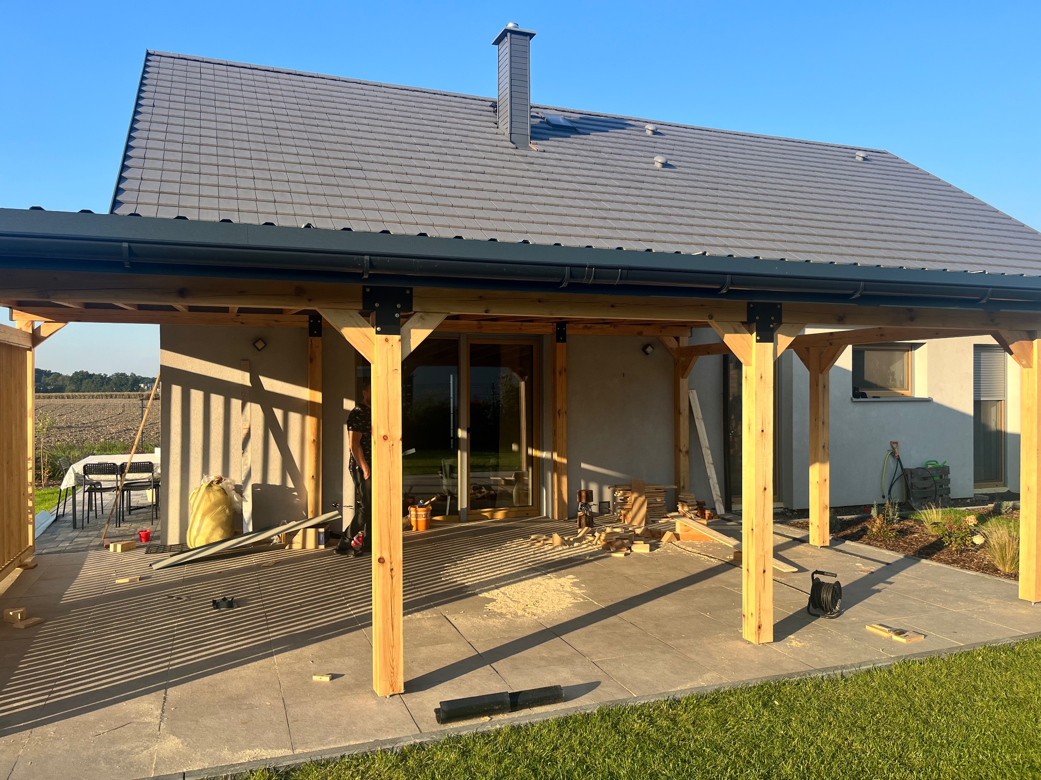 Drewniana pergola w trakcie budowy, widoczne słupy i zadaszenie, narzędzia pracy, dom w tle, słoneczny dzień. Elementy konstrukcyjne w surowym drewnie.