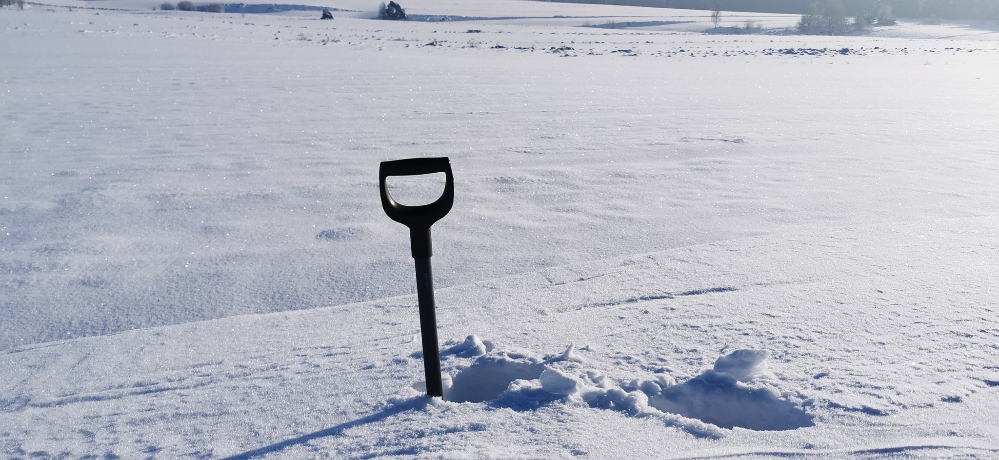 Czarna łopata wbita w śnieżnobiały, zmrożony grunt, z wykopanym obok dołem, w tle rozległe pole i pojedyncze drzewa.