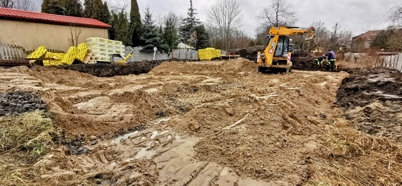 Żółta koparko-ładowarka JCB na terenie budowy, wyrównywanie terenu pod budowę, widoczne pryzmy piasku i błota, w tle materiały izolacyjne i budynek z czerwonym dachem.