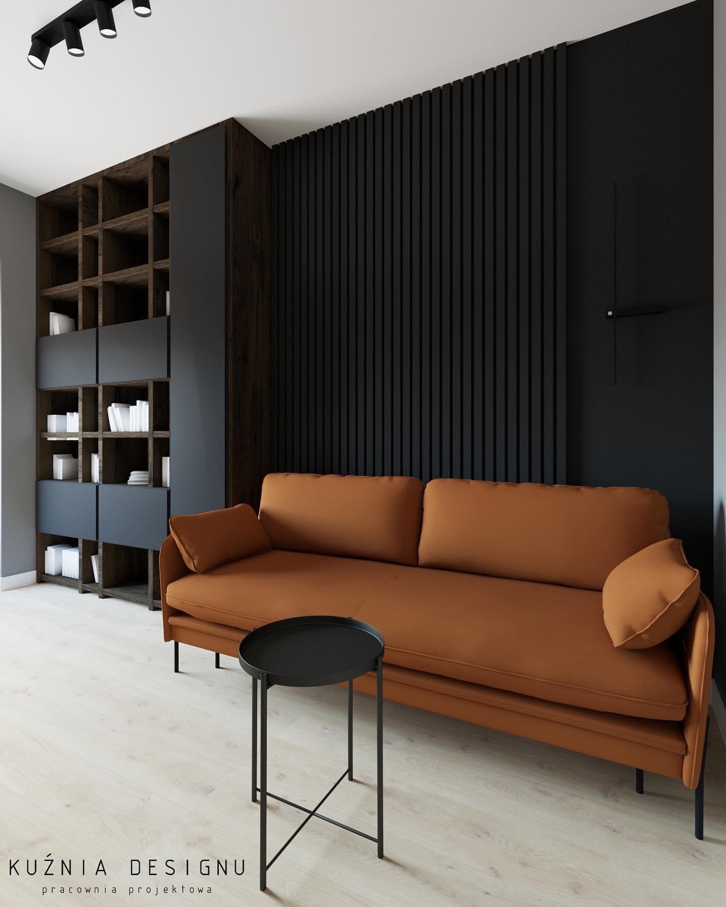 Minimalistyczny salon z terakotową sofą, czarnymi pionowymi panelami ściennymi i drewnianym regałem z szarymi elementami, oświetlony reflektorami sufitowymi.